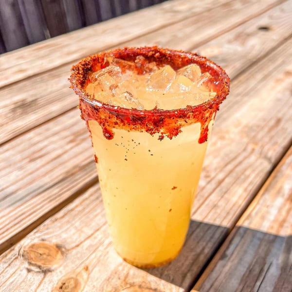 A yellow drink with ice and a red, spicy rim sits on a wooden picnic table.