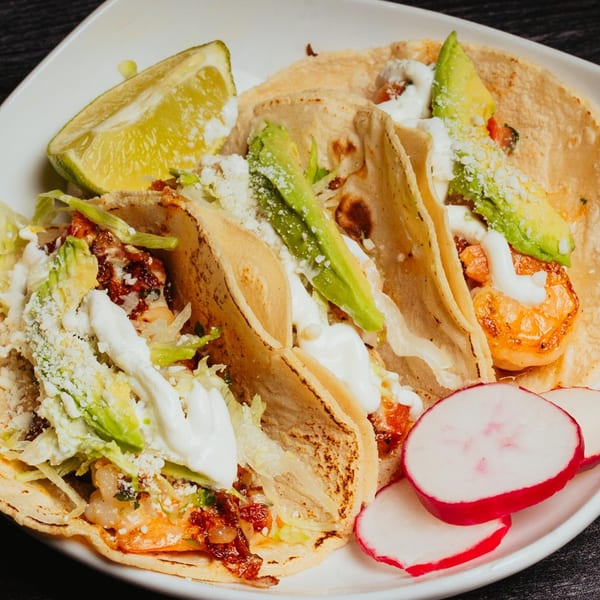 Three shrimp tacos with avocado, sour cream, and cheese, served with a lime wedge and radishes.