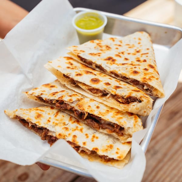 Four quesadilla slices filled with meat and cheese, served with green sauce in a metal tray.