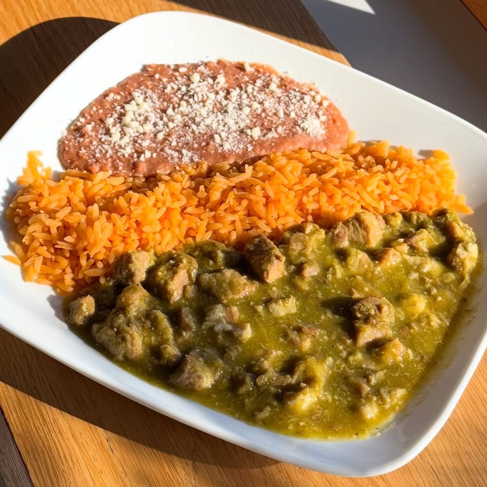 Pork chile verde with orange rice and refried beans topped with cheese on a white plate.