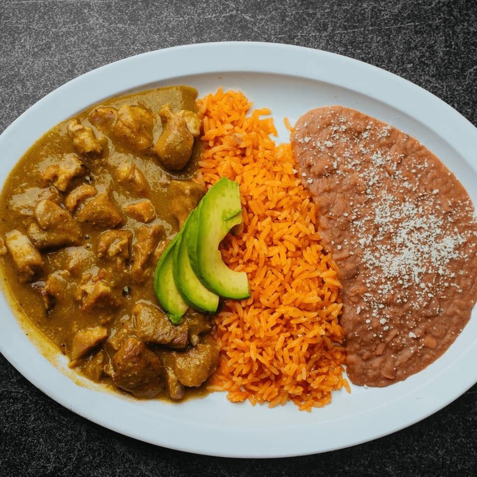 Pork in green chile, orange rice, and refried beans with avocado slices on a white plate.