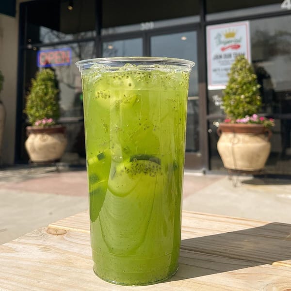 Green drink with chia seeds and fruit chunks in a plastic cup on a wooden table outside.