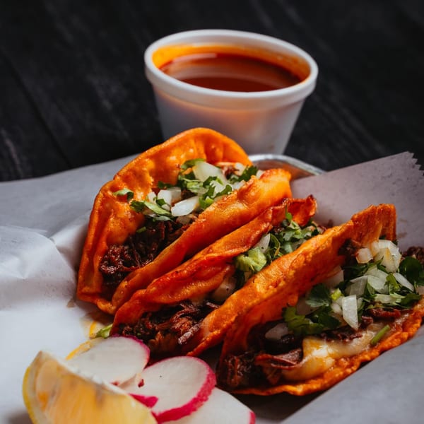 Three birria tacos with cilantro, onions, and cheese, served with a side of consommé, lemon, and radishes.