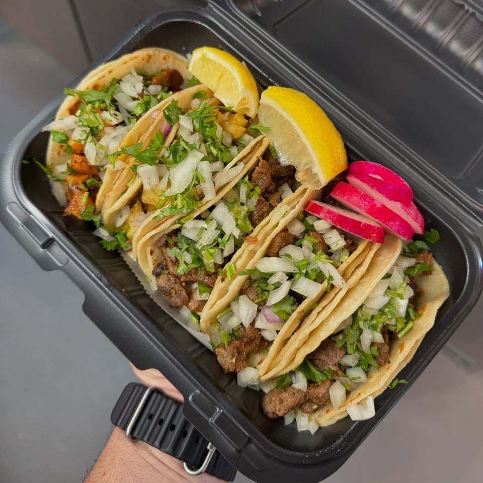 A hand holds a black takeout container filled with five street tacos, garnished with lemon and radishes.