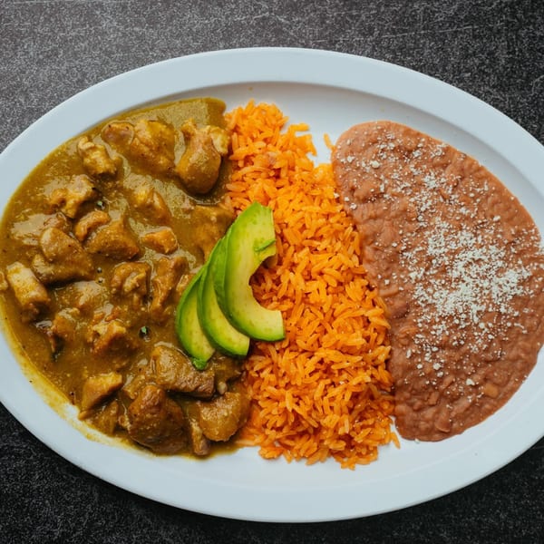 Pork in green chile sauce with orange rice, refried beans, and avocado slices on a white plate.