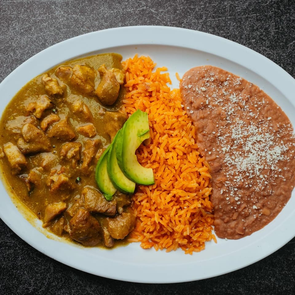 Pork in green chile sauce with orange rice, refried beans, and avocado slices on a white plate.