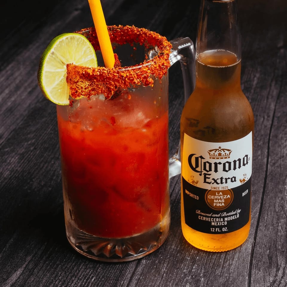 A michelada in a mug with a lime and a Corona Extra beer bottle on a dark wooden surface.