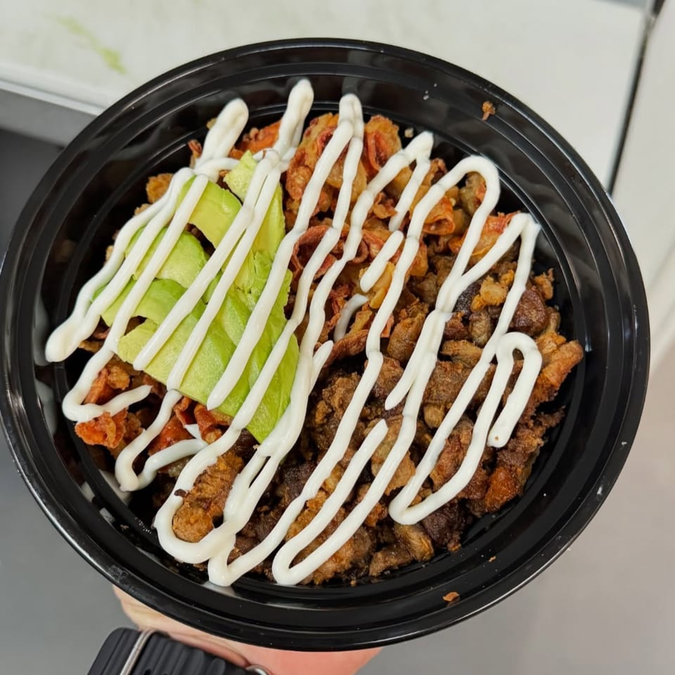 Crispy pork belly and chicharrones with avocado slices and a creamy white sauce in a black bowl.
